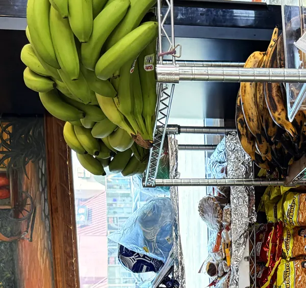 Green and ripe plantains hanging above shelves of snacks and red beans.